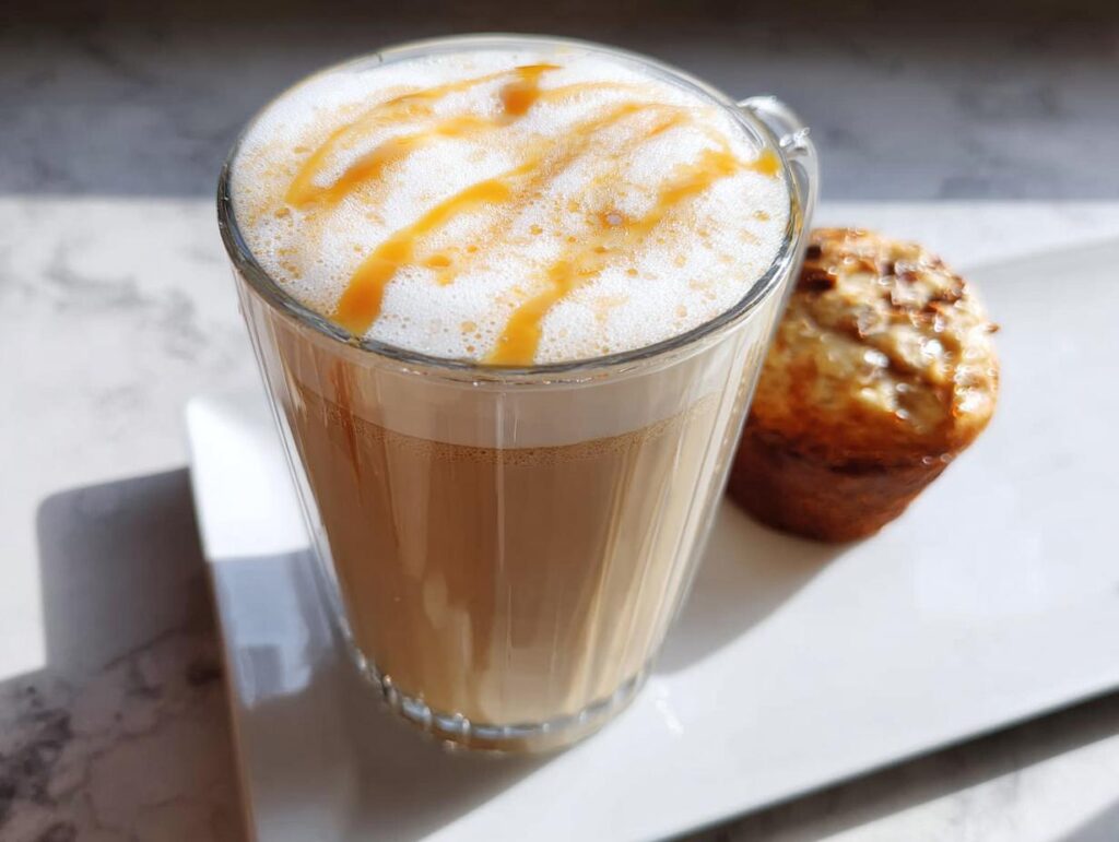 A tall glass of Caramel Macchiato Style At Home coffee with foamy milk and caramel drizzle, next to a muffin.