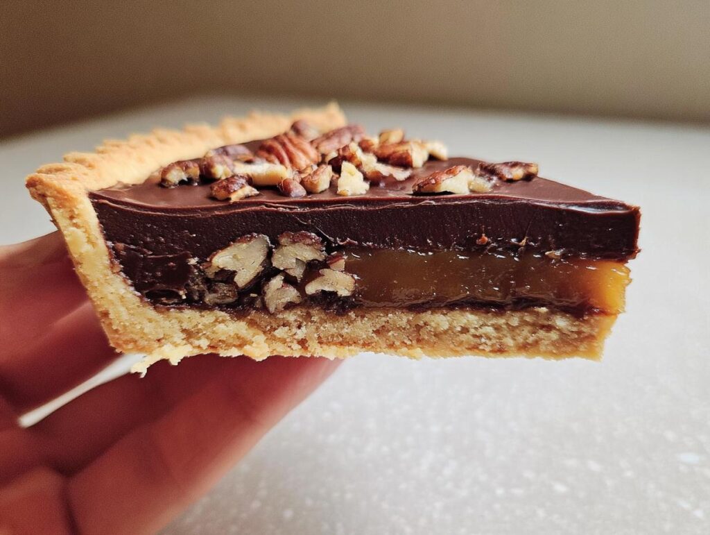 A slice of rich chocolate tart with toasted pecans, showing layers of chocolate ganache and caramel.