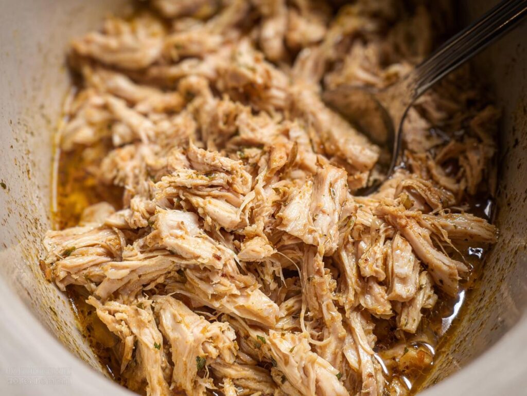 Close-up of tender, shredded chicken thighs cooked in a slow cooker, glistening with juices and herbs.