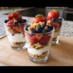 Three glasses filled with Yogurt Parfaits With Fruit And Crunch, layered with yogurt, granola, strawberries, and blueberries.